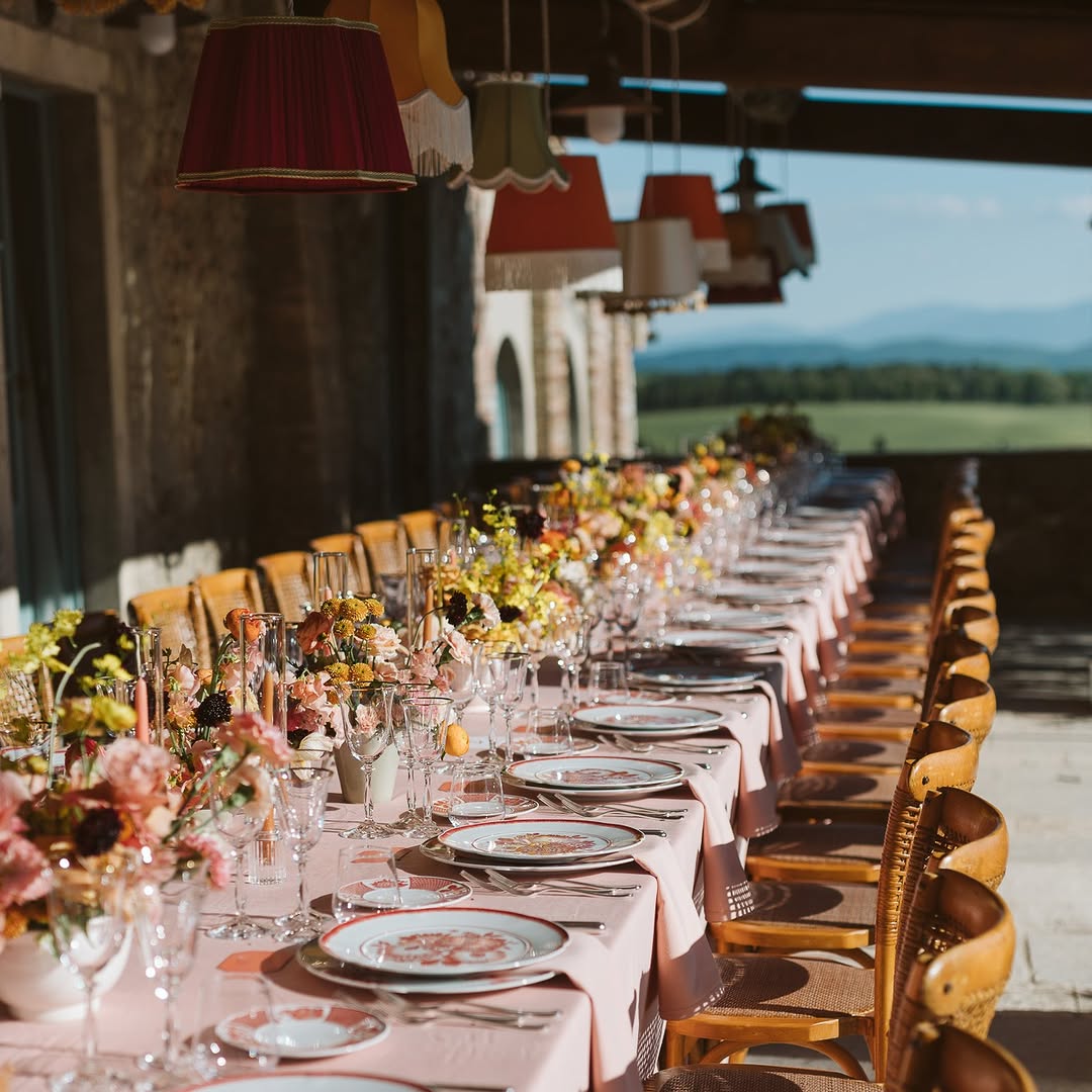 A scenic long-table wedding reception setup at Borgo Pignano, one of the top Tuscany wedding venues, with blush pink linens, vintage floral china, and vibrant spring florals, set on a sunlit stone terrace overlooking the Tuscan hills.