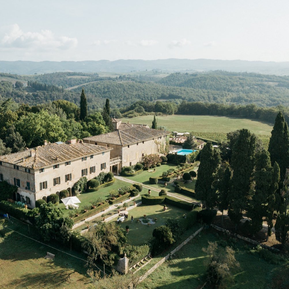 Aerial view of Borgo Stomennano, a breathtaking Tuscany wedding venue nestled amidst rolling vineyards and manicured gardens, showcasing historic stone buildings and a large landscaped lawn perfect for elegant outdoor weddings.