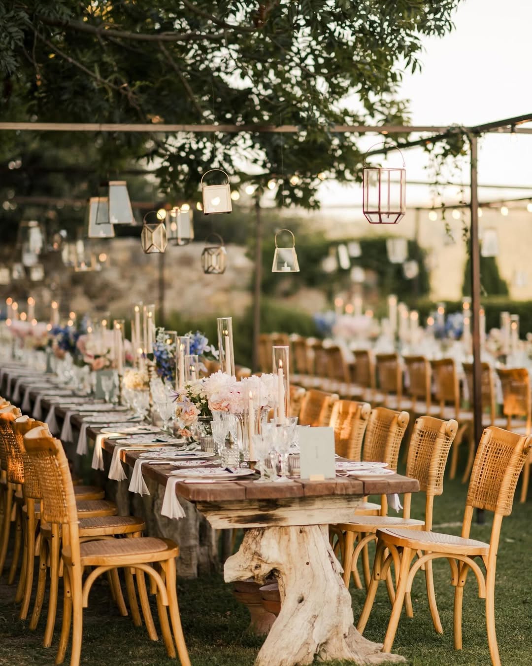 Rustic wedding dinner setup at Castello di Vicarello, a charming Tuscany wedding venue, featuring wooden tables, rattan chairs, and hanging lanterns under a canopy of treesβideal for a whimsical garden celebration.