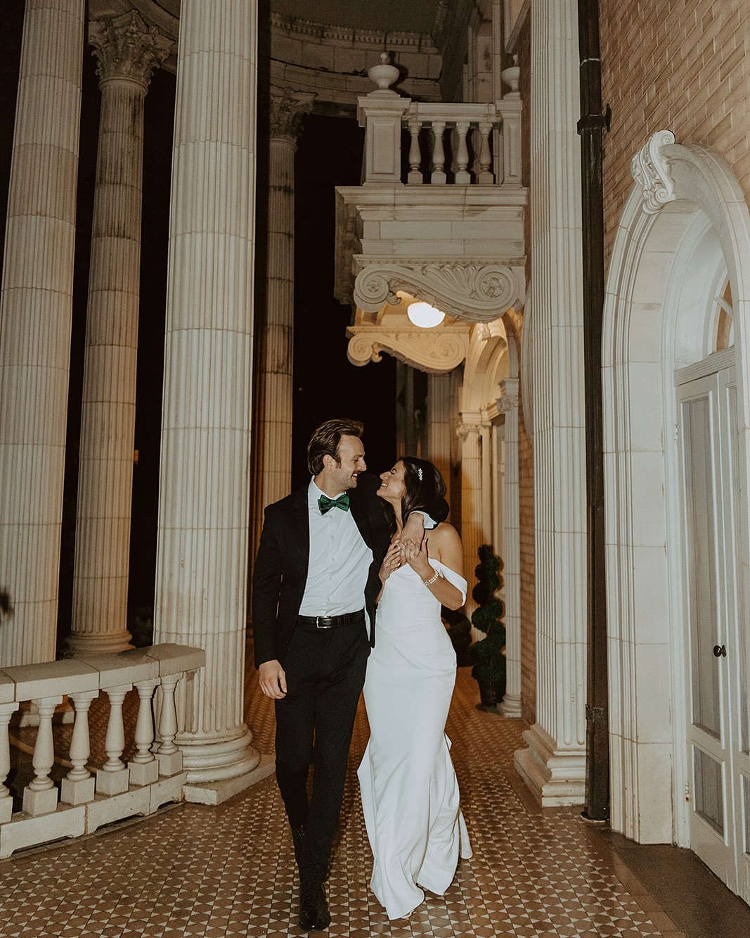 Elegant bride and groom walking at night in front of the historic columns and intricate architecture of the Grant-Humphreys Mansion β one of the most timeless Denver wedding venues.