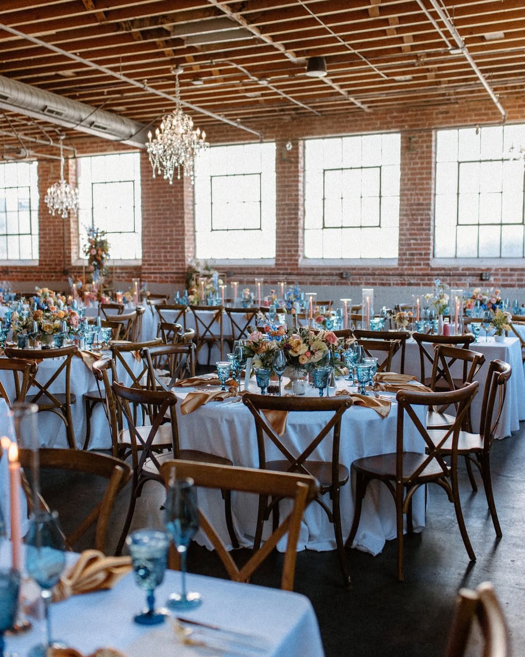 Industrial-chic wedding reception at Moss Denver with exposed brick walls, crystal chandeliers, and blue and gold table settings β a modern gem in the heart of Denver wedding venues.