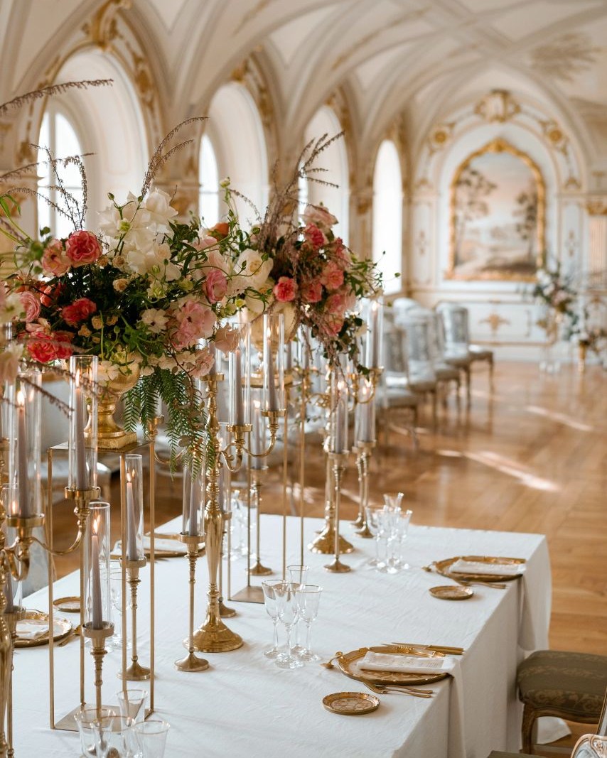 Lavish ballroom reception at Relais La Corte dei Papi, an opulent Tuscany wedding venue with vaulted white-and-gold ceilings, floor-to-ceiling arched windows, and tall gold candelabras adorned with pink and white florals.