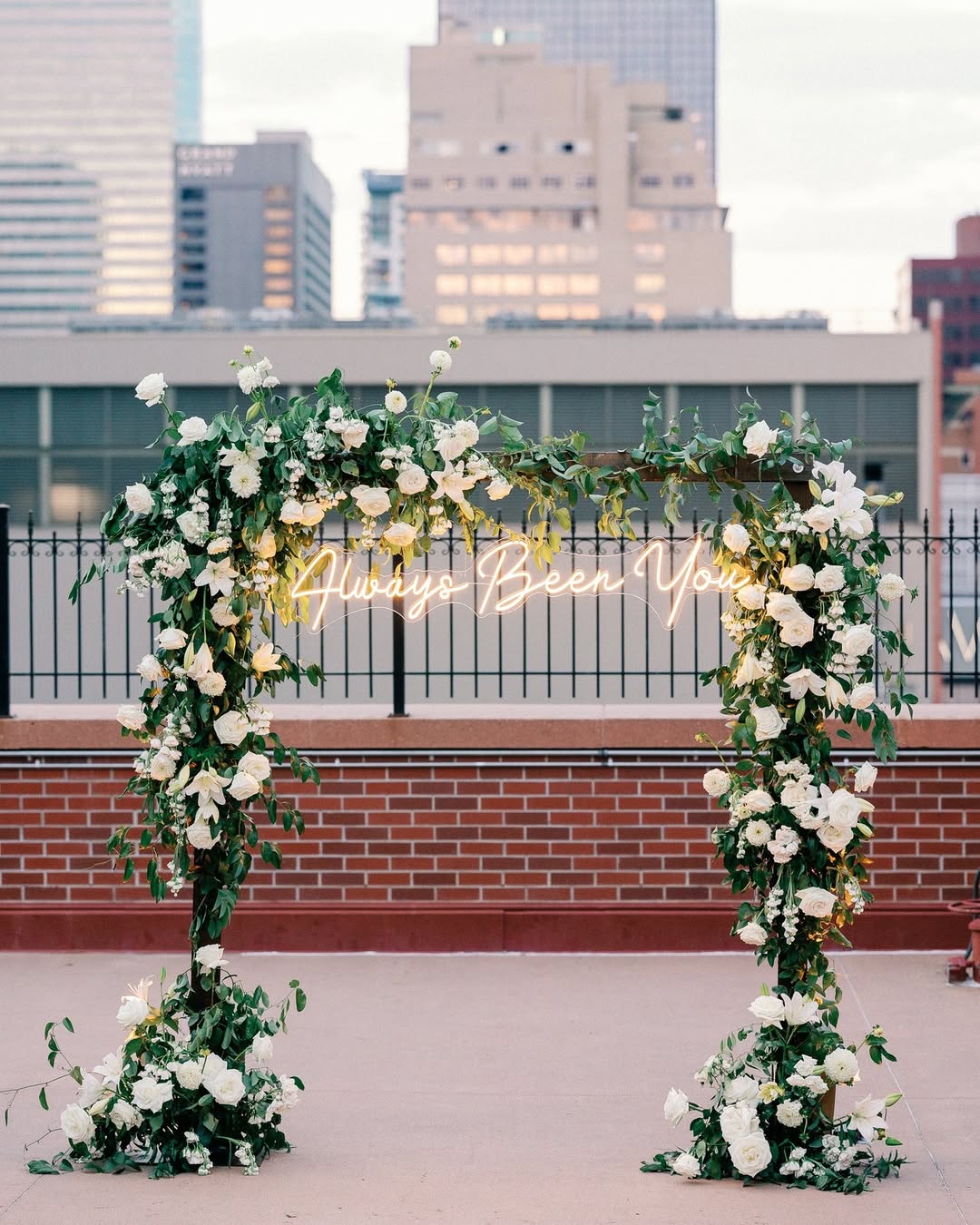 Rooftop wedding ceremony with lush floral arch and 'Always Been You' neon sign against the city skyline at The Denver Athletic Club β an iconic city-view Denver wedding venue.
