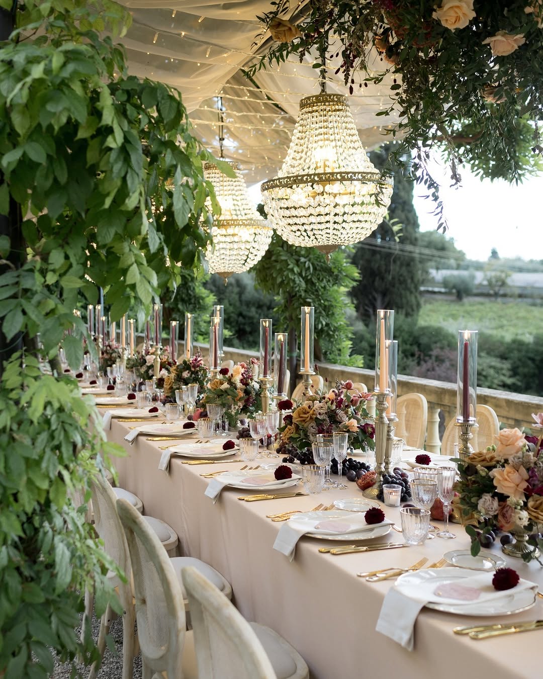 Intimate outdoor wedding table setup at Villa Le Fontanelle, a luxury Tuscany wedding venue, styled with cream linens, antique chandeliers, lush green foliage, and romantic candlelit dΓ©cor under a canopy of vines.