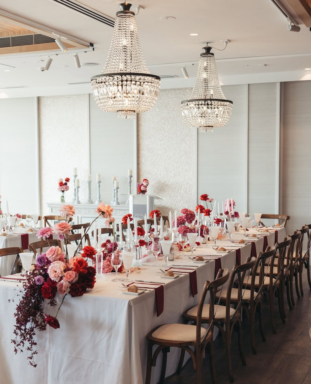 A long banquet table decorated with pink, red, and blush florals is set beneath sparkling chandeliers at Zest Waterfront Venues, a top sydney wedding venue offering stylish indoor spaces with waterfront elegance.